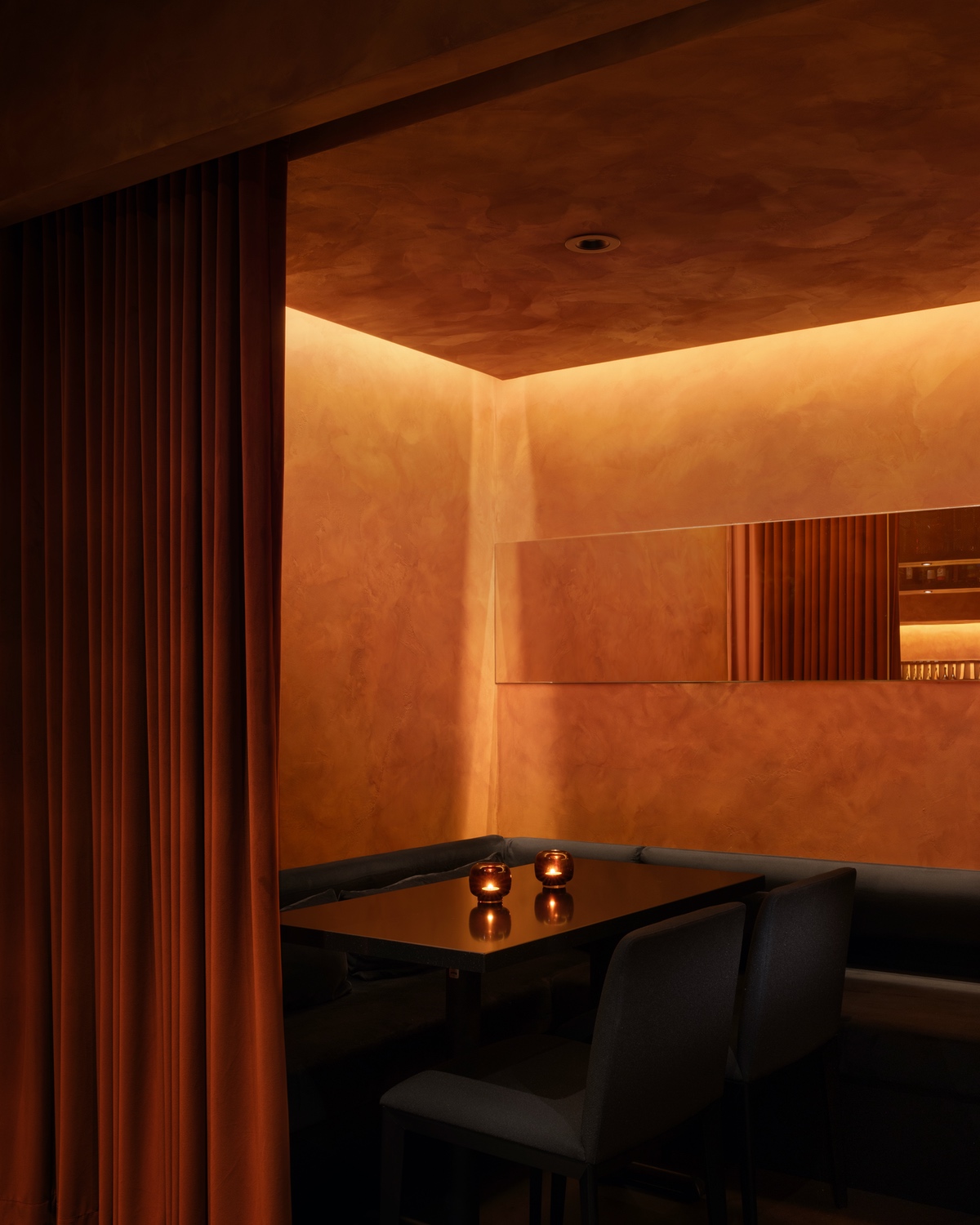 Ama restaurant by Vancouver design studio &Daughters is modeled after Blade Runner 2049. A black table sits in a dining nook closed off with orange velvet curtains. Behind it are a golden mirror and venetian plaster walls soaked in a golden glow.