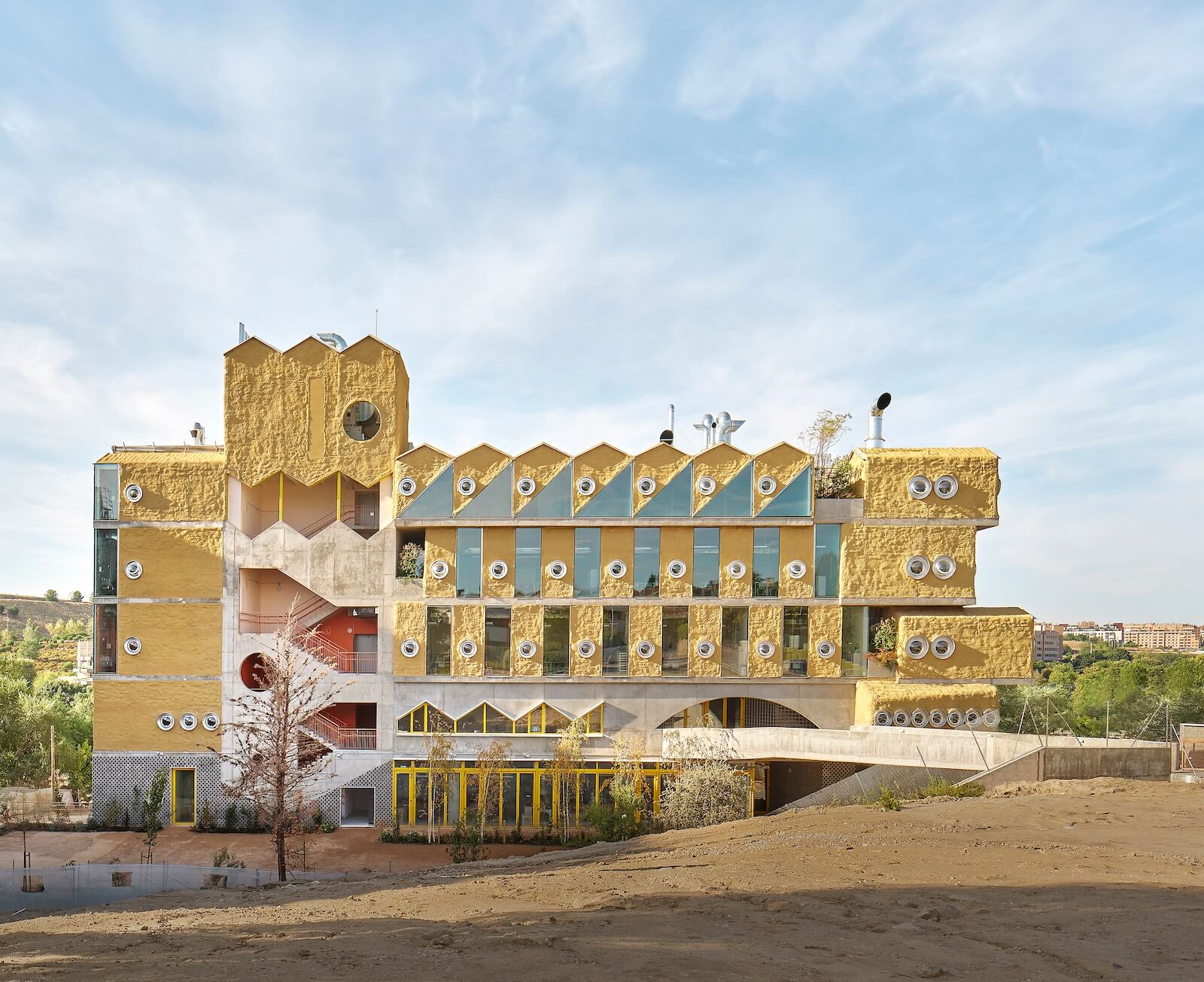 A zigzagging roofline and porthole windows animate the Reggio School in Madrid by Andres Jaque. Its yellow cork cladding is expected to attract colonies of lichen and fungi, while its gardens are designed to entice wildlife including birds, bats and butterflies.