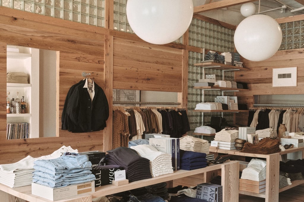 An image of a Buck Mason store design in Oak Brook, Illinois by Norman Kelley, featuring an oakwood shell structure built inside of a storefront. A black chore coat hangs on the side wall of the structure, while a window-like cutout looks out to the white periphery space where shelves display additional clothing.