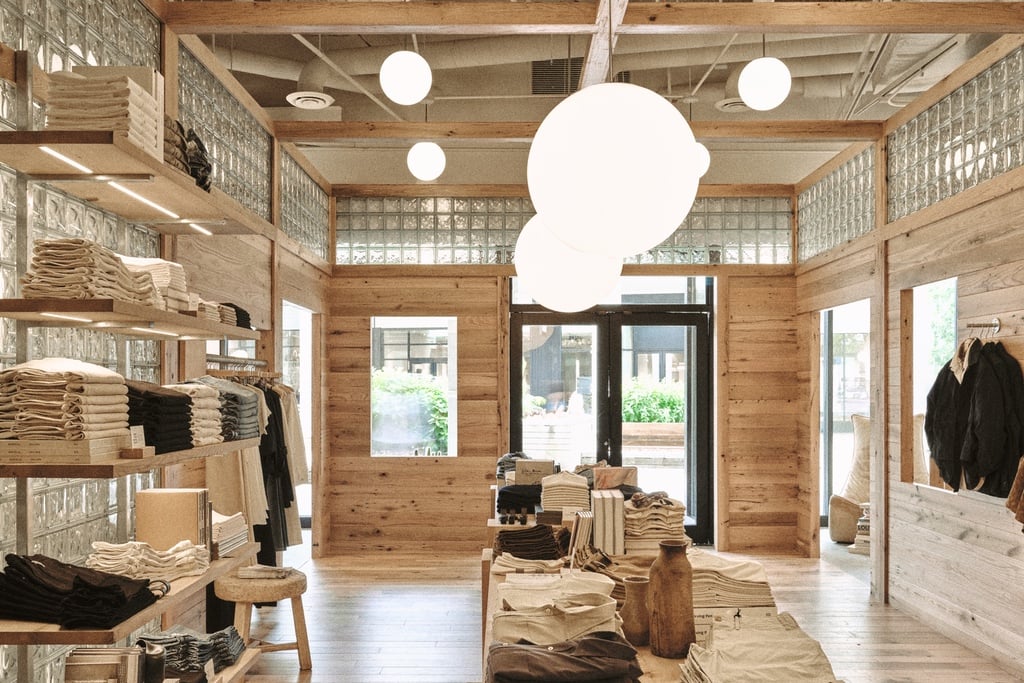 An image of a Buck Mason store design in Oak Brook, Illinois by Norman Kelley, featuring an oakwood shell structure built inside of a storefront. The top of the wooden structure is lined in glass brick and orb-style lights hang above tables of clothing in khaki and grey tones.