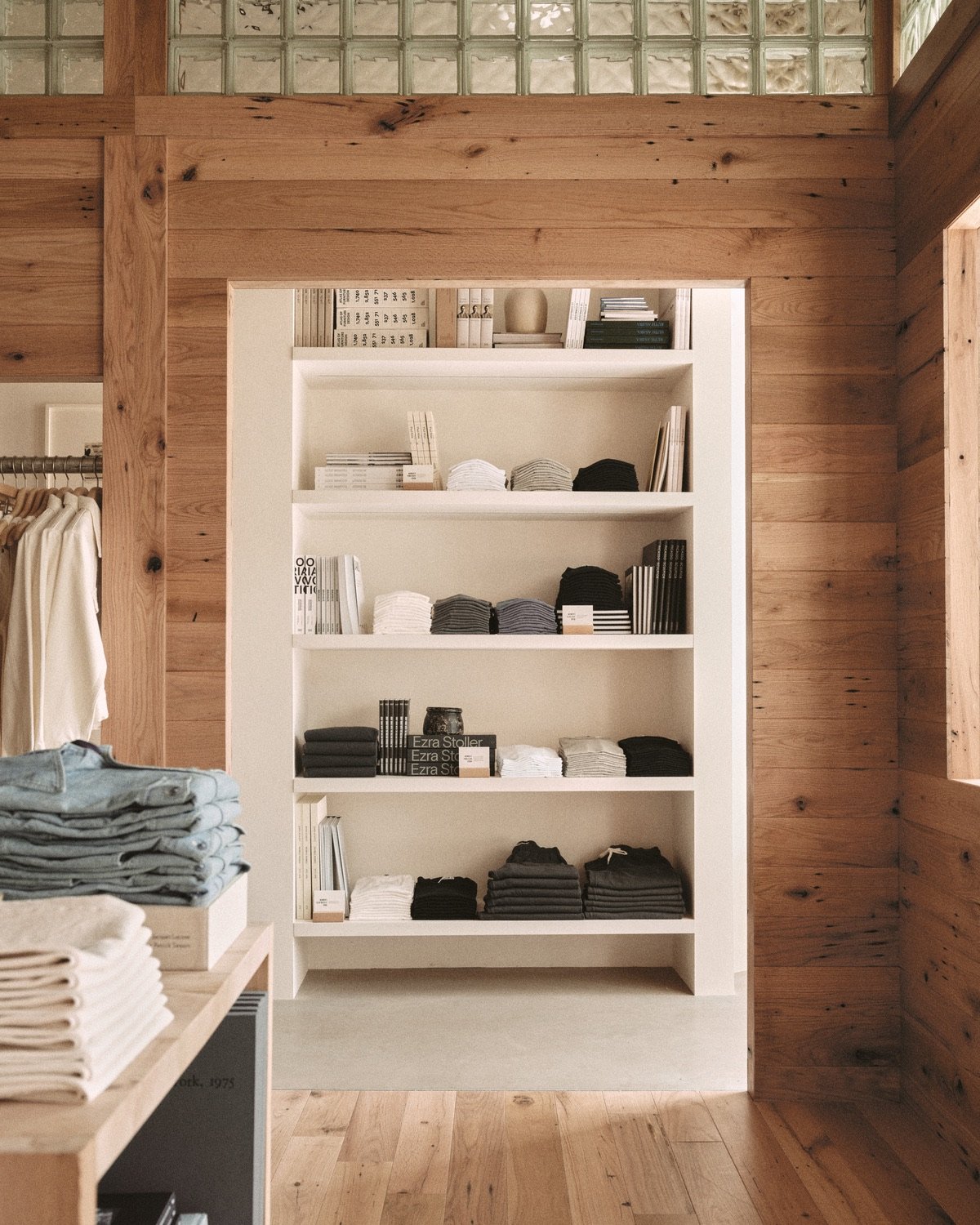 An image of a Buck Mason store design in Oak Brook, Illinois by Norman Kelley, featuring an oakwood shell structure featuring a clerestory row of glass bricks. A doorway cutout looks out from this interior shop-within-shop to the perimeter space, where shelves display stacks of clothing.