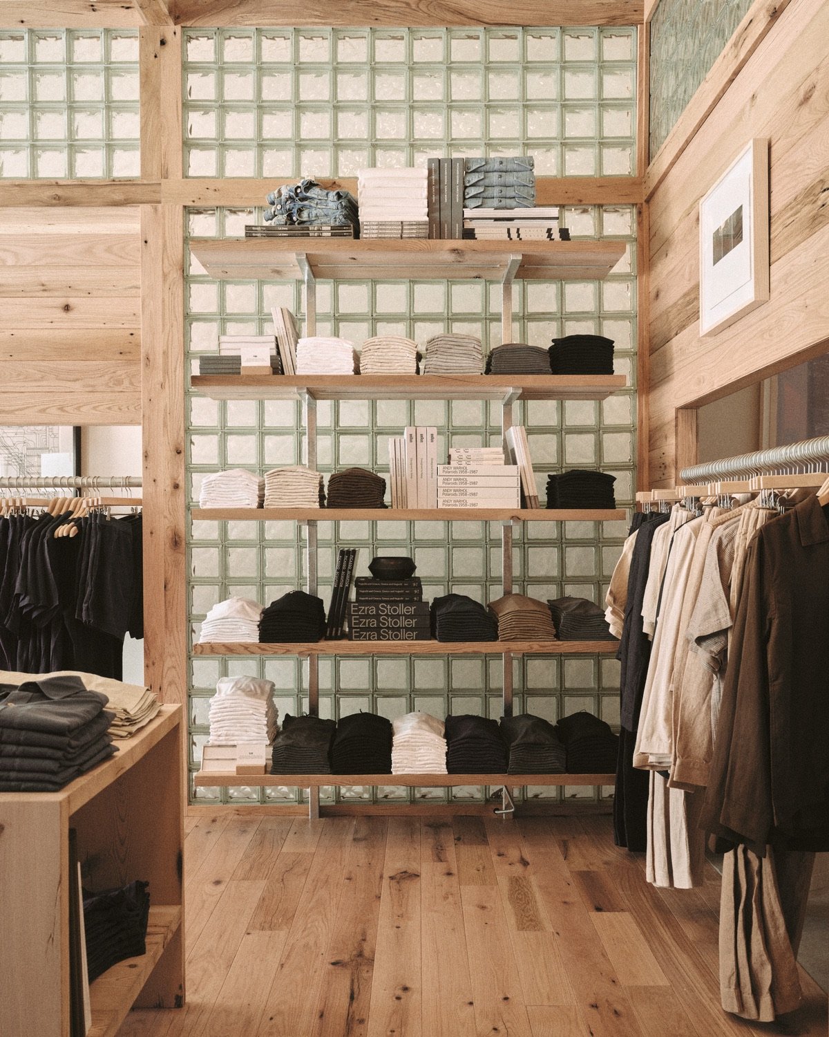 An image of a Buck Mason store design in Oak Brook, Illinois by Norman Kelley, featuring an oakwood shell structure. Here, a wall clad in glass bricks features wooden shelves displaying stacks of sweaters.