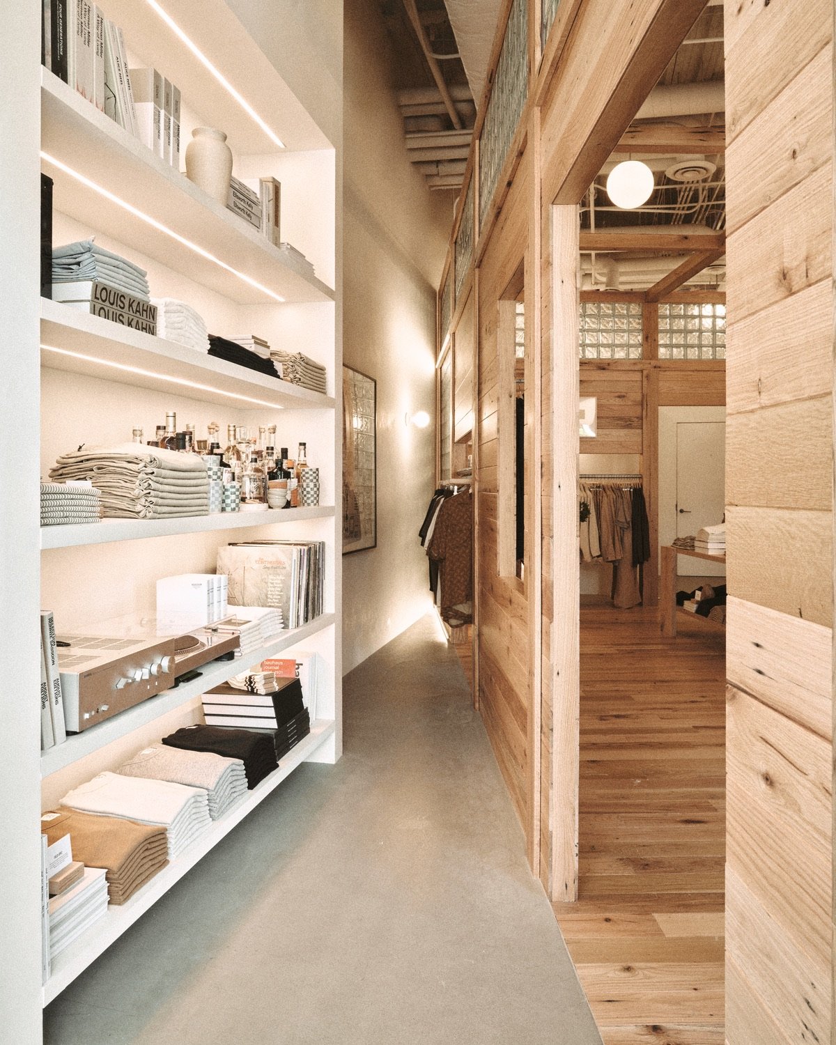 An image of a Buck Mason store design in Oak Brook, Illinois by Norman Kelley, featuring an oakwood shell structure built inside of a storefront. The shell structure is built at an angle to the existing walls, forming a triangular nook in the perimeter of the space. To the left, white shelving on the outer walls displays a selection of stacked t-shirts. A doorway to the white steps into the interior shop-within-shop.