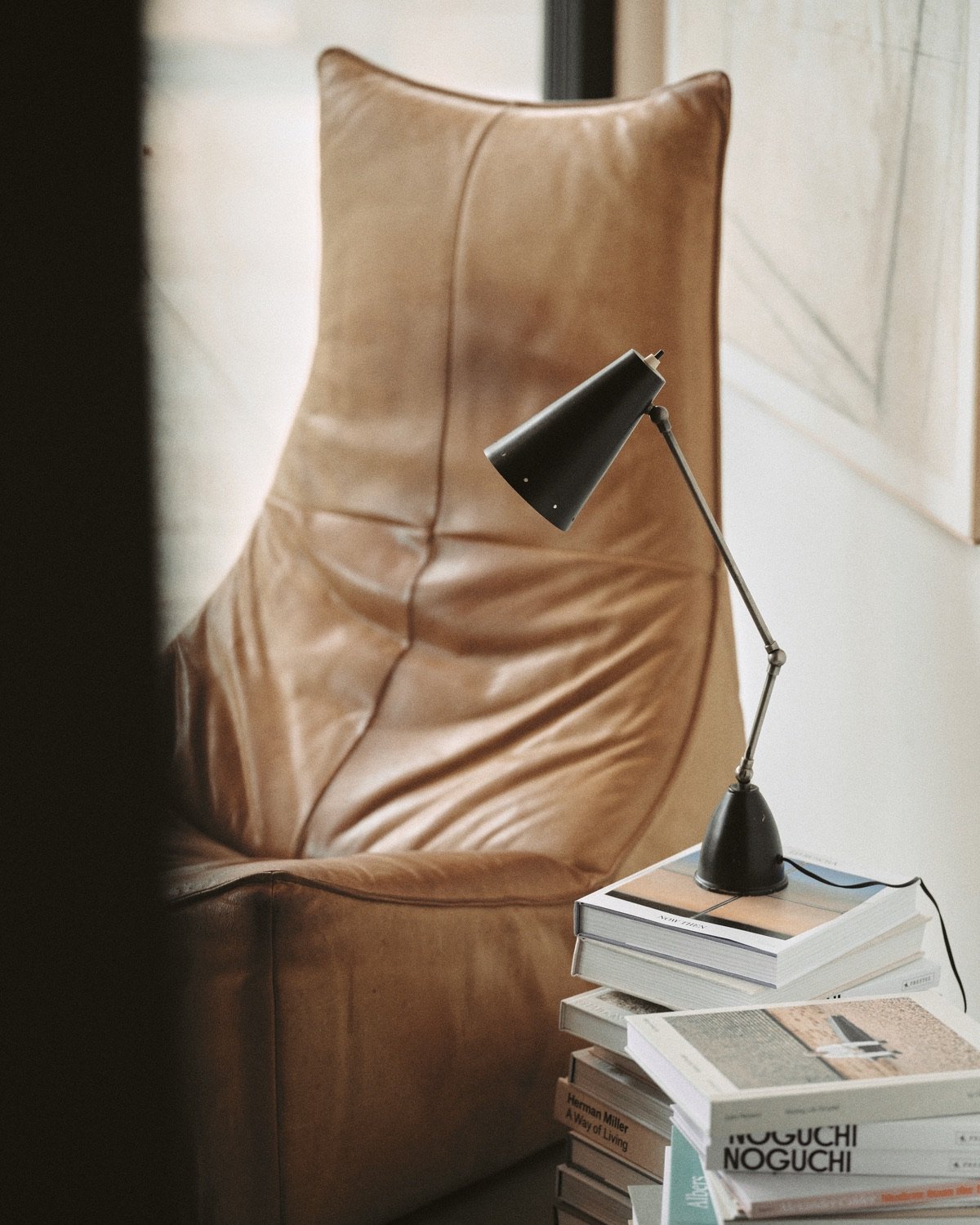 A black table lamp rests on a stack of design books next to a vintage brown leather chair.