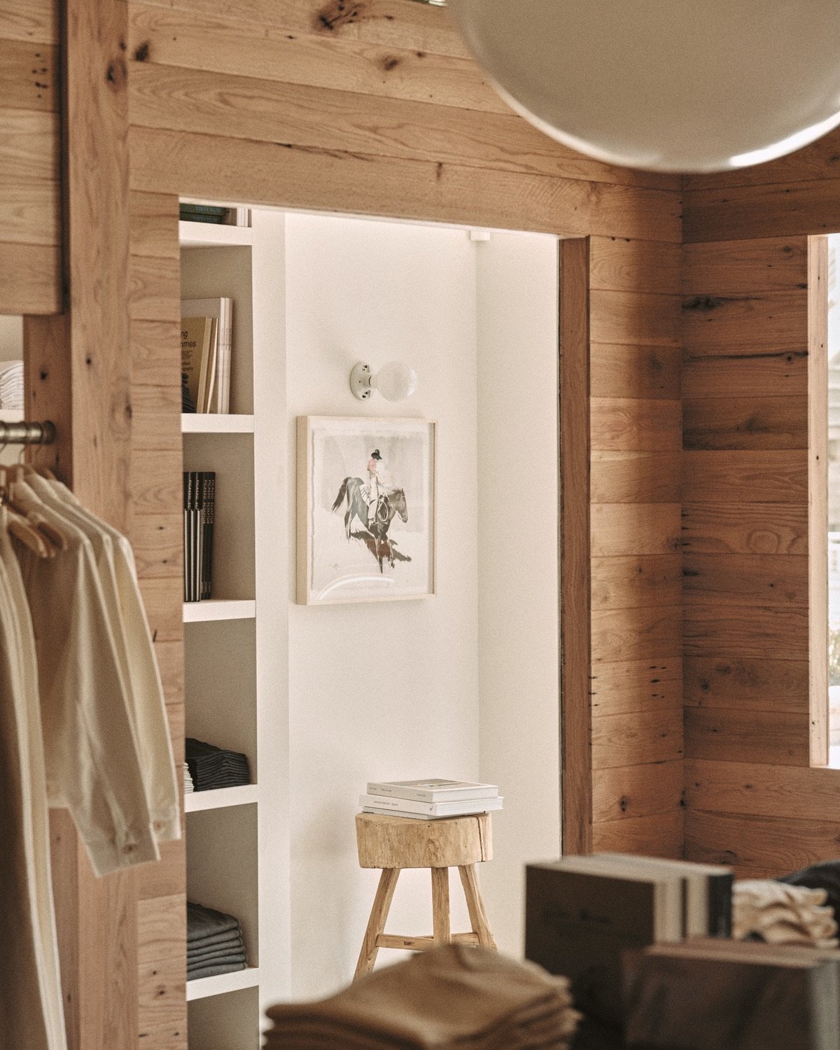 An image of a Buck Mason store design in Oak Brook, Illinois by Norman Kelley, featuring an oakwood shell structure built inside of a storefront. A doorway cutout looks out from this interior shop-within-shop to the perimeter space, where a framed photo of a cowboy riding a bucking bronco hangs on the white wall.