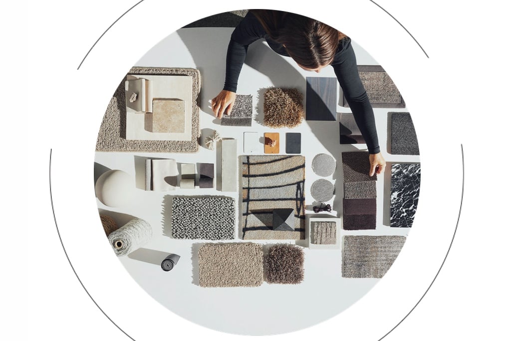 A woman leans over a table featuring fabric samples, highlighting the sustainable materials used by Age Carpets. On top of the image, thin black lines form a circle, highlighting an example of circular design in the furniture industry.