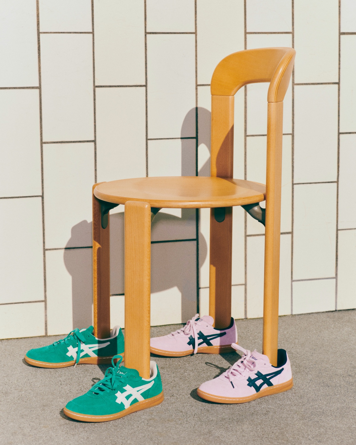 Two pairs of Asics sneakers (one green with white stripes, one pink with dark blue stripes) made in collaboration with design brand Hay, with the legs of a wooden Hay chair resting inside of them.