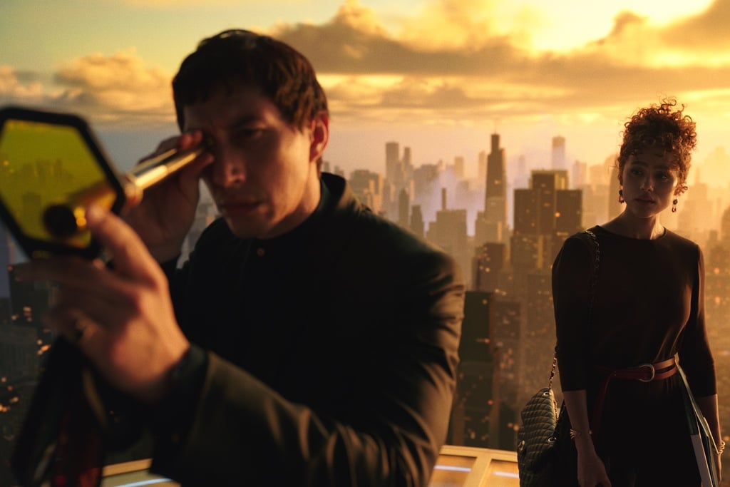 A still from the Francis Ford Coppola movie Megalopolis shows Adam Driver and Nathalie Emmanuel in front of an orange-tinted skyline with the architecture of a modern city. Driver is holding a telescope-like object with a hexagonal yellow glass chip at the end.