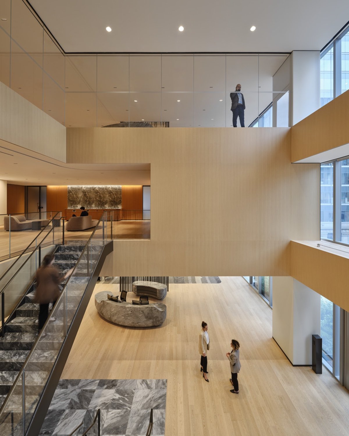 A view of an open, three-storey space in the Scotiabank North office designed by KPMB Architects in Toronto with light wood flooring and a marble staircase connecting the first two floors. A man stands in a glass window looking into the atrium from the third floor.