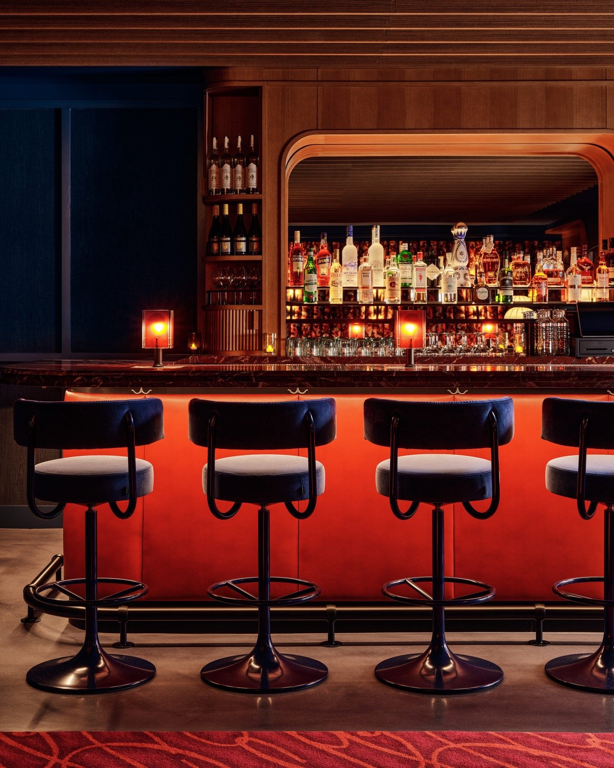 A row of blue velvet bar stools sit in front of a red bar at So&So's designed by Goodrich. Bottles of liquor sit against a mirror framed in wood.