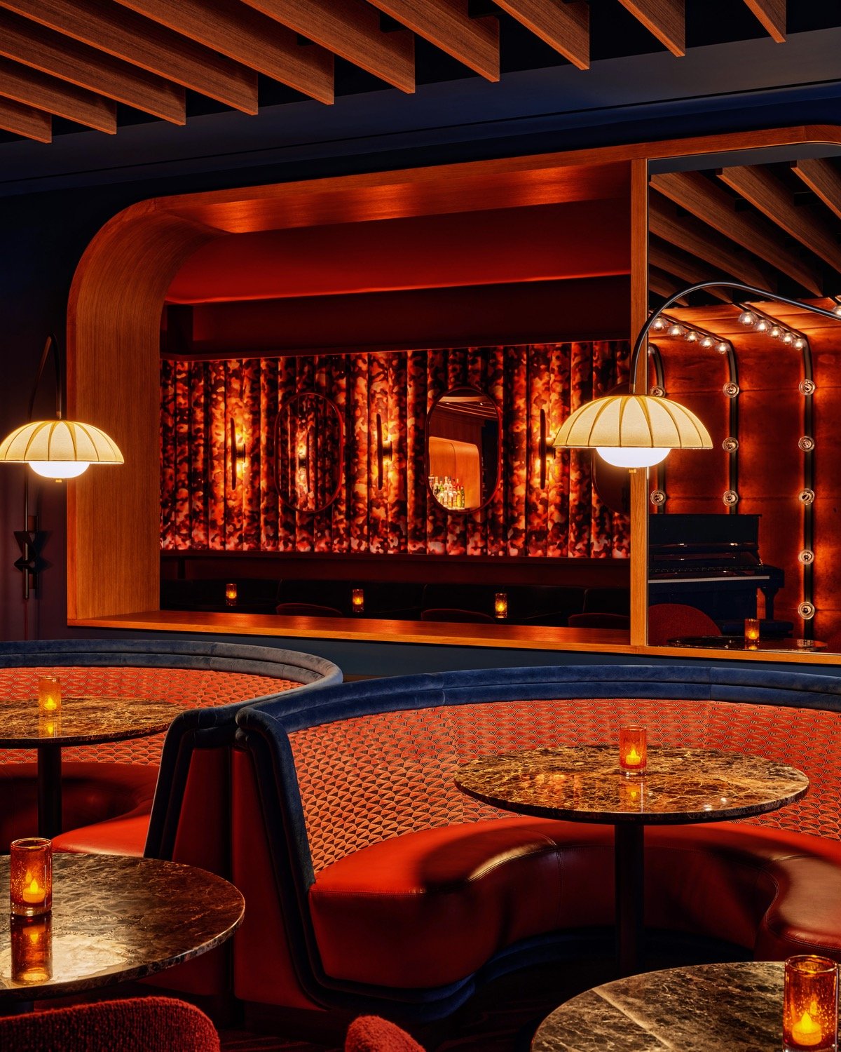 A view of the red banquette seating at New York piano bar So&So's designed by Goodrich. Arced lamps extend out from the wall over circular marble cocktail tables.