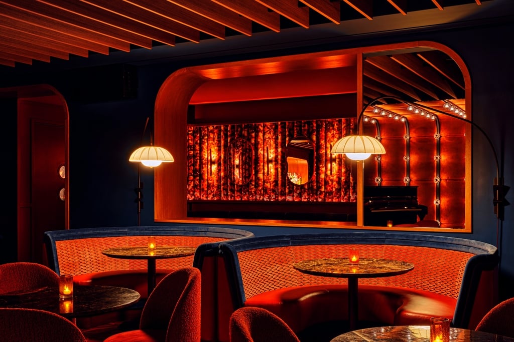 A view of the red banquette seating at New York piano bar So&So's designed by Goodrich. Arced lamps extend out from the wall over circular marble cocktail tables.