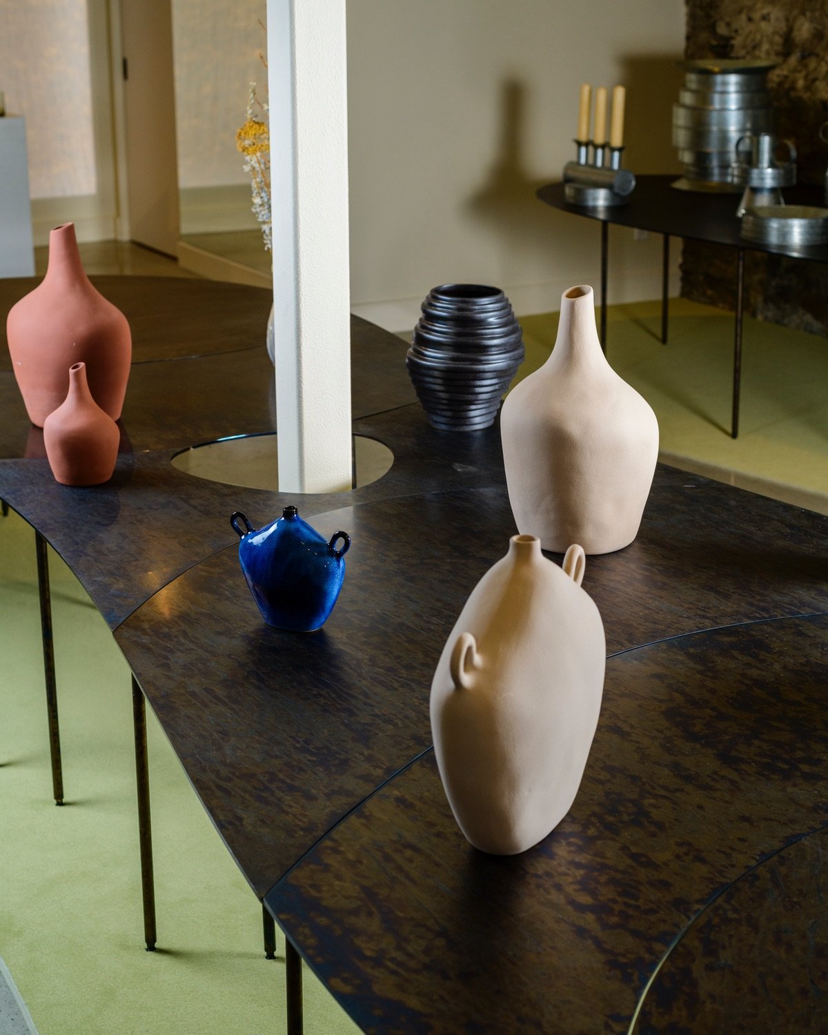 A selection of sculptural vases sit on a rounded black steel table in front of a natural stone wall at the Montreal store 207 Ouest. A circular cutout in the middle of the table allows a column to pass through.