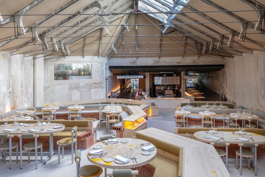 The dining room at Tramo restaurant in Madrid, featuring tiered levels of dining tables created from industrial materials like steel and plywood. Above is an overhead skylight and a kitchen area waits at the bottom.